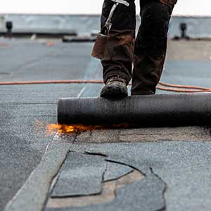 Worker installing roofing membrane with torch, illustrating building wear and repair tied to property obsolescence in real estate