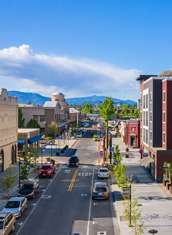 Aerial view of Market Center in Downtown Redding, CA, showcasing a bustling commercial area with modern buildings, vibrant storefronts, and pedestrian-friendly streets. The center is surrounded by landscaped plazas, wide sidewalks, and nearby parking lots. The image captures the lively atmosphere of the downtown area, with people walking and shopping, cars moving through the streets, and scenic mountain views in the background. The architecture blends contemporary design with the charm of the local area, creating a hub for community activity.